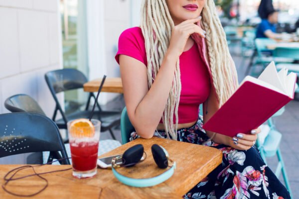 blonde-unusual-woman-with-dreadlocks-sitting-cafe-street-holding-notebook-enjoying-free-time-wearing-bright-pants-with-tropical-print
